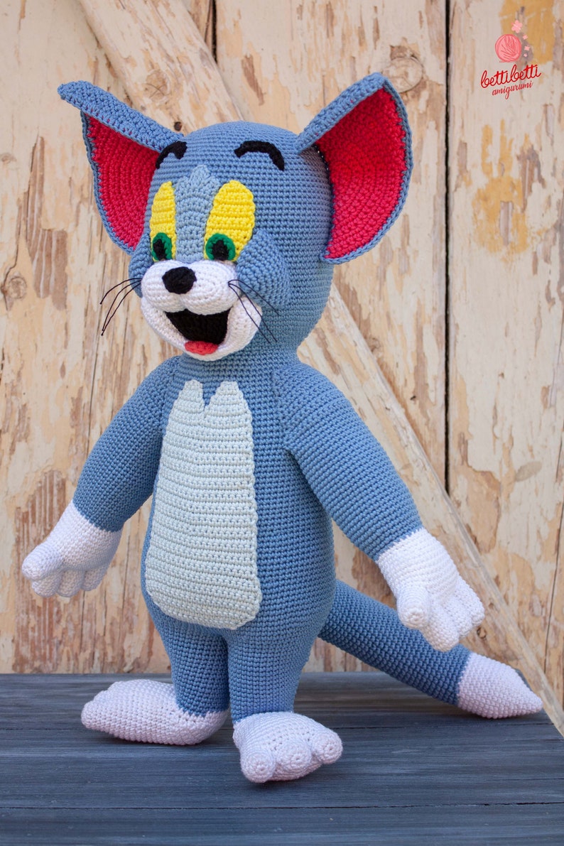 May include: A crocheted Tom cat plush toy with blue fur, white paws, and a red and white face. The toy is standing on a wooden surface.