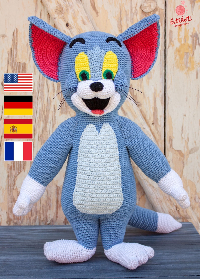 May include: A crocheted Tom and Jerry character, Tom the cat, is blue with white paws and a red inner ear. The cat is smiling with a white mouth and a red tongue. The cat has yellow eyes with green pupils. The cat is standing on a wooden surface.