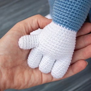 May include: A white crocheted hand with four fingers, held by a person's hand. The crocheted hand is attached to a blue and white crocheted arm.