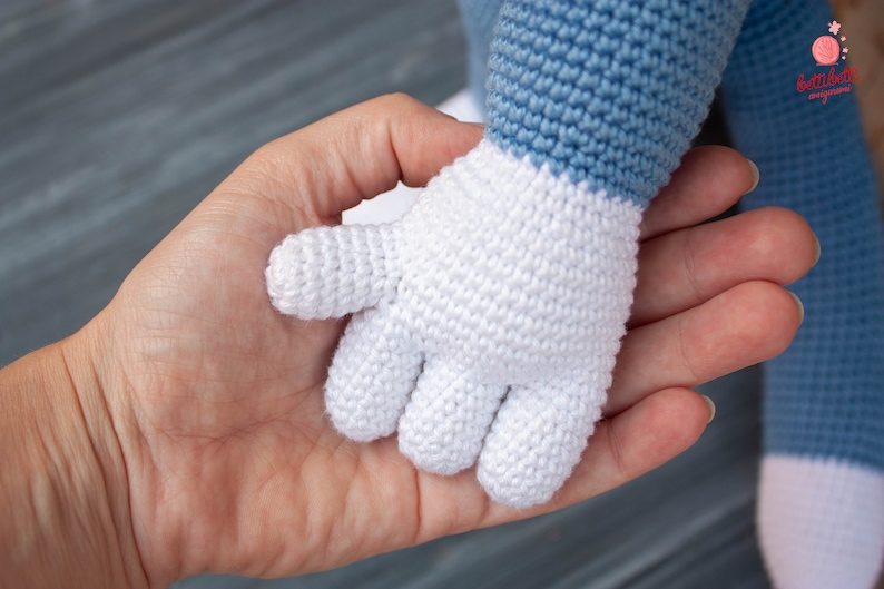 May include: A white crocheted hand with four fingers, held by a person's hand. The crocheted hand is attached to a blue and white crocheted arm.