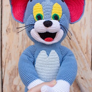 May include: A crocheted Tom and Jerry character, Tom the cat, is blue with white accents. He has red ears, green eyes, and a yellow nose. He is smiling with his mouth open.