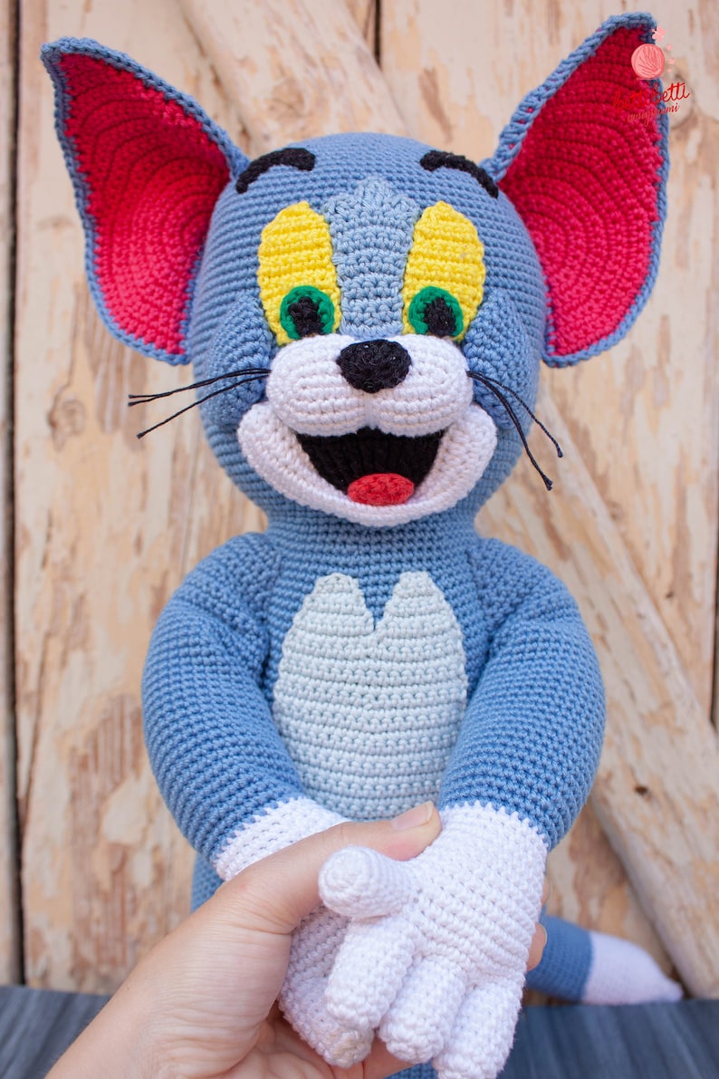May include: A crocheted Tom and Jerry character, Tom the cat, is blue with white accents. He has red ears, green eyes, and a yellow nose. He is smiling with his mouth open.