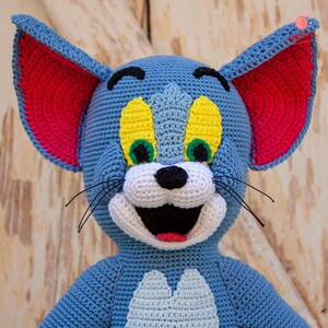 May include: A crocheted plush toy of Tom the cat from the cartoon Tom and Jerry. The toy is blue with yellow eyes, a white nose, and a red mouth. The ears are pink and the whiskers are black.
