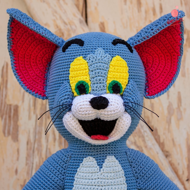 May include: A crocheted plush toy of Tom the cat from the cartoon Tom and Jerry. The toy is blue with yellow eyes, a white nose, and a red mouth. The ears are pink and the whiskers are black.