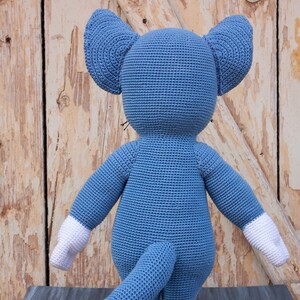 May include: A blue crocheted cat toy with white crocheted feet and tail. The cat is standing on a gray wooden surface.