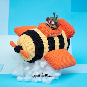 May include: A crocheted bee-shaped aeroplane with orange wings and a yellow and black striped body. The pilot is a small crocheted figure with a green hat and orange scarf. The aeroplane is resting on a cloud. The image includes the text @bettibettiami.