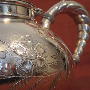 May include: Close-up of a silver teapot with floral engravings. The teapot has a twisted handle and a lid. The surface is shiny, reflecting light and highlighting the detailed floral patterns.