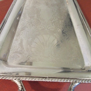May include: A rectangular silver serving tray with a decorative floral pattern and a rope-like border. The tray has curved edges and is supported by ornate legs. The tray is likely made of metal.