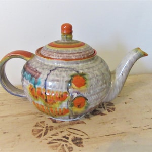 May include: A white ceramic teapot with a lid and a handle. The teapot is decorated with orange, yellow, and green floral designs. The teapot is sitting on a light wood surface.