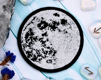 May include: A black and white moon patch with a white background and a black moon design. The patch is surrounded by small white stones with gold runes on them.