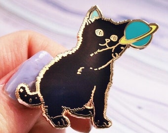 Puede incluir: Pin esmaltado de gato negro con ribete dorado, sosteniendo un planeta turquesa con un anillo amarillo. El gato tiene detalles azules en las orejas. Un accesorio divertido para chaquetas, bolsos o sombreros.