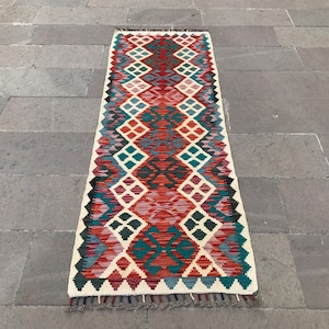 Puede incluir: Una alfombra larga y rectangular con un patrón geométrico en tonos de rojo, azul, verde y blanco. La alfombra tiene flecos en ambos extremos.