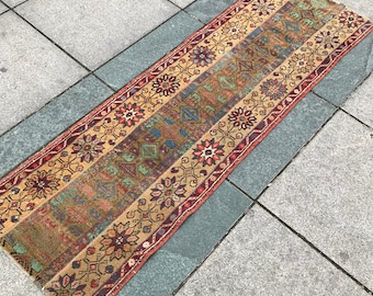 May include: A vintage Turkish runner rug with a colorful geometric pattern in shades of red, blue, green, and beige. The rug has a fringed edge and is laid out on a gray tiled floor.