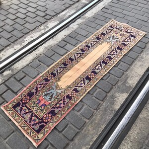 May include: A vintage runner rug with a beige background and a colorful geometric pattern. The rug has a dark purple border with pink and green accents.