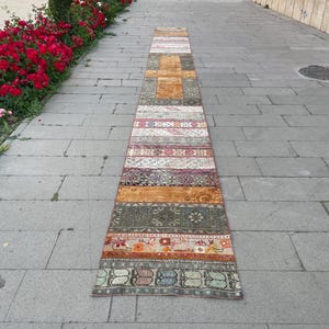 Könnte beinhalten: Ein langer, schmaler Läufer mit einem Patchwork-Design, das aus verschiedenen rechteckigen Abschnitten besteht. Der Teppich zeigt eine vielfältige Farbpalette mit Tönen in Orange, Olivgrün, Dunkelrot, Rosa und gedämpftem Grau sowie komplizierten geometrischen und floralen Mustern auf der gesamten Textiloberfläche.