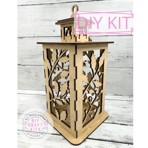 May include: A laser-cut wooden lantern kit with a rectangular shape and a decorative holly leaf design. The lantern has a handle on top and the text "DIY KIT" is visible. The base has a triangular shape. The lantern is made of light brown wood.