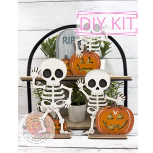 May include: A Halloween DIY kit featuring wooden skeleton figures and carved orange pumpkins. The skeletons are waving, and the pumpkins have carved faces. The kit includes a tombstone and the text "DIY KIT".