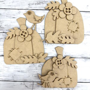 May include: Three unfinished wooden pumpkin cutouts with a bird and leaves. The pumpkins are layered and have a button in the center of the flower.