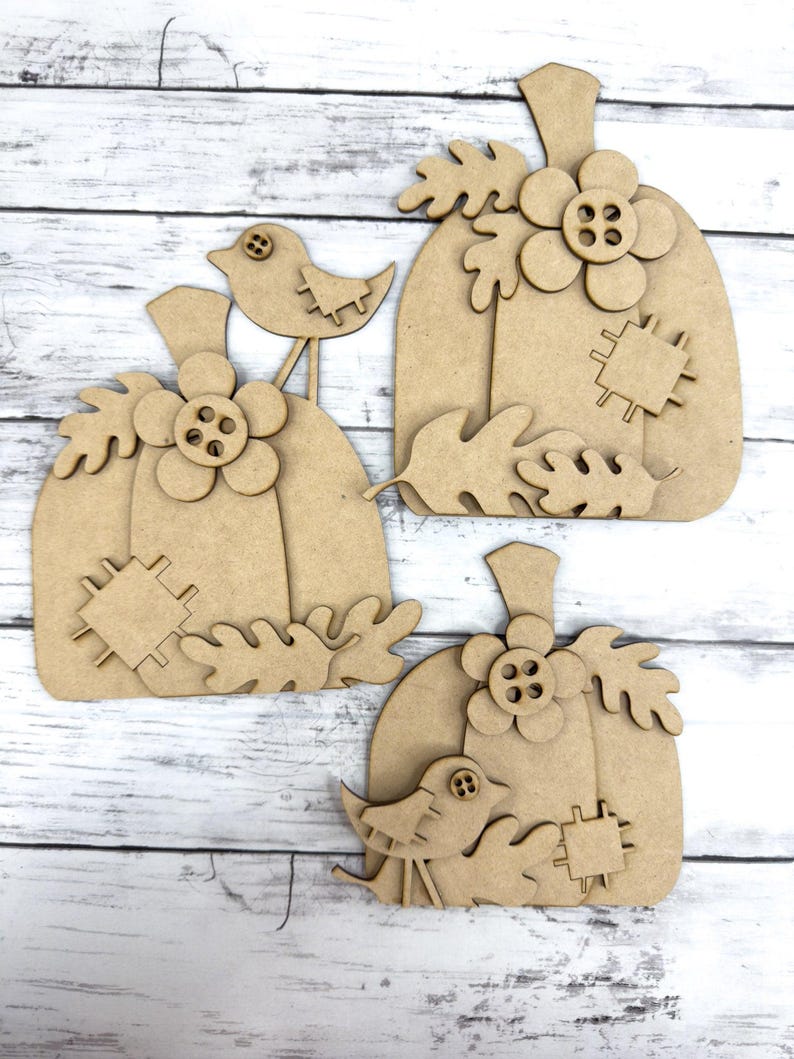 May include: Three unfinished wooden pumpkin cutouts with a bird and leaves. The pumpkins are layered and have a button in the center of the flower.