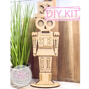 May include: A wooden DIY kit of a nutcracker soldier. The kit is made of light-colored wood and features a skull-shaped head, a traditional hat, and a detailed uniform. The words "DIY KIT" are visible in pink text. The kit is displayed with a potted plant.