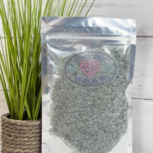 May include: A clear, resealable bag filled with small, gray, irregularly shaped pieces. The bag has a label with text and a heart design. A potted green plant with long blades sits to the left of the bag.