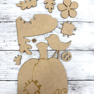 May include: A wooden craft kit featuring a pumpkin, leaves, a bird, a flower, and a button. The pieces are unfinished and ready to be painted or stained.