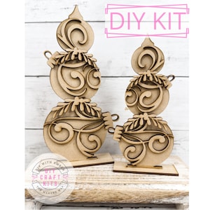 May include: DIY craft kit featuring two wooden ornaments shaped like Christmas ornaments with intricate designs. The ornaments are stacked on top of each other and have a base. The kit is labeled "DIY Kit" and "DIY with Amber Craft Kits The Makers Map".