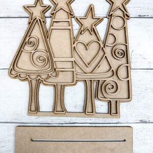 Op de afbeelding: Vier onafgewerkte houten kerstboomversieringen met verschillende ontwerpen. Elke boom heeft een ster bovenop en een basis om te staan. Een simpel houten bord is inbegrepen.