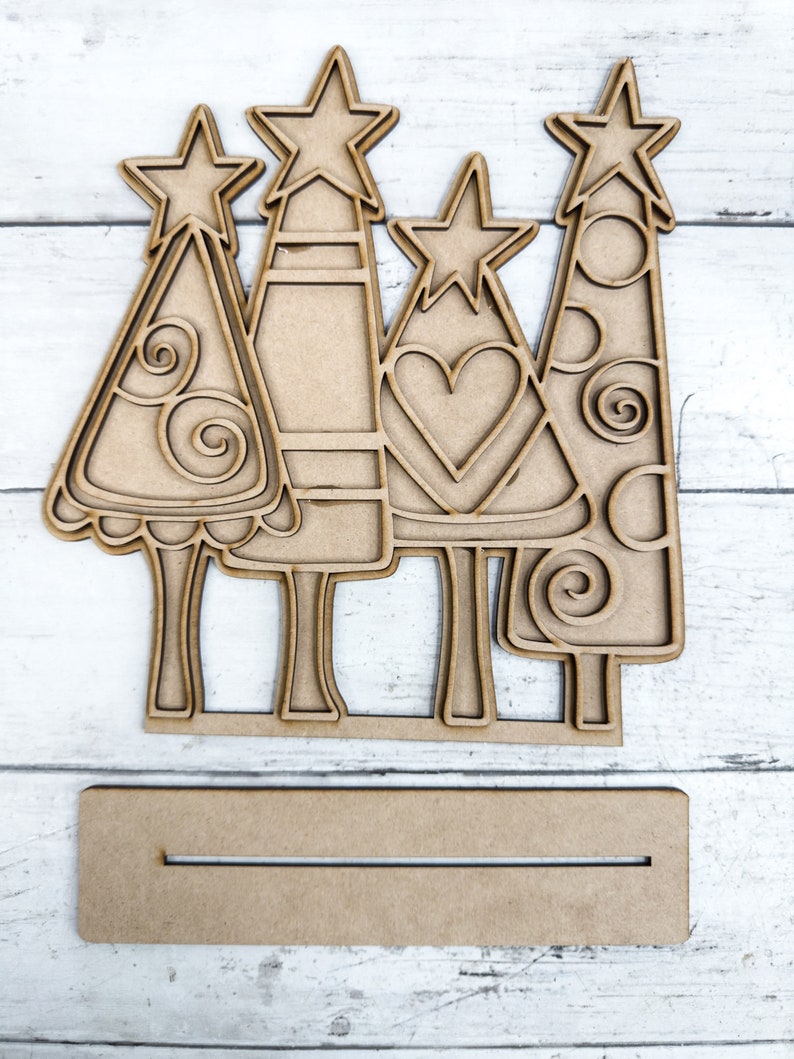 Op de afbeelding: Vier onafgewerkte houten kerstboomversieringen met verschillende ontwerpen. Elke boom heeft een ster bovenop en een basis om te staan. Een simpel houten bord is inbegrepen.