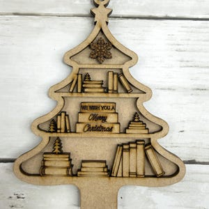 May include: Wooden Christmas tree ornament with shelves filled with books and small trees. The ornament has a snowflake and the text "WE WISH YOU A Merry Christmas". The ornament is made of light brown wood.