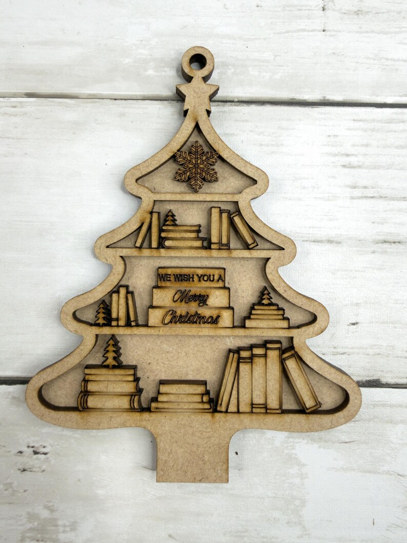 May include: Wooden Christmas tree ornament with shelves filled with books and small trees. The ornament has a snowflake and the text "WE WISH YOU A Merry Christmas". The ornament is made of light brown wood.