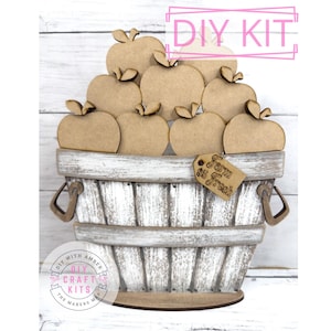 May include: DIY craft kit featuring a wooden basket filled with apples. The basket is whitewashed and has a rustic look. The apples are unpainted and ready to be decorated. The kit includes a sign that says "Farm $5 Fresh".