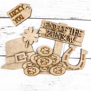 May include: Wooden craft supplies with a St. Patrick's Day theme. The set includes a leprechaun hat, a pot of gold coins, a horseshoe, a shamrock, and a sign that reads "End of the Rainbow". There is also a tag that reads "Lucky You".