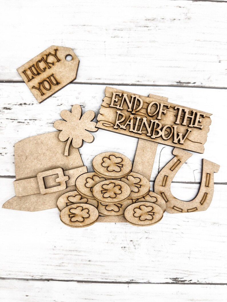 May include: Wooden craft supplies with a St. Patrick's Day theme. The set includes a leprechaun hat, a pot of gold coins, a horseshoe, a shamrock, and a sign that reads "End of the Rainbow". There is also a tag that reads "Lucky You".