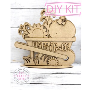 May include: Wooden craft kit featuring a wheelbarrow filled with a pumpkin, sunflowers, and fall foliage. The wheelbarrow has the words "Happy Fall" written on it. The kit is made by DIY with Amber Craft Kits.