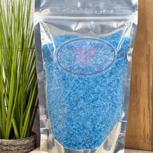 May include: A clear, silver-lined bag filled with bright blue crystals. The bag has a clear front and a silver back. The text "Stay with Amber" is printed on a blue oval label. The bag is on a wooden surface.