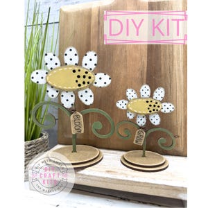 May include: DIY flower kit with two wooden flower-shaped decorations. The flowers have white petals with black polka dots and a yellow center. The stems are green with a swirl design and a wooden base. The text "DIY KIT" is in a pink frame.