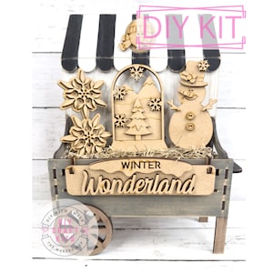May include: A wooden DIY kit featuring a winter wonderland theme. The kit includes a cart with a striped awning, snowflakes, a snow globe with mountains and a tree, and a snowman. The words "WINTER Wonderland" are displayed.
