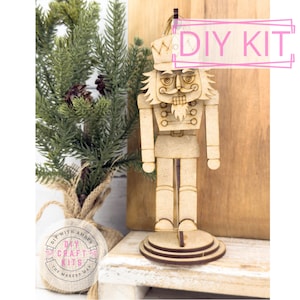 May include: A wooden DIY kit of a nutcracker soldier with a detailed design. The kit includes a base and a hanging loop. The text "DIY KIT" is visible. A small evergreen tree is in the background.