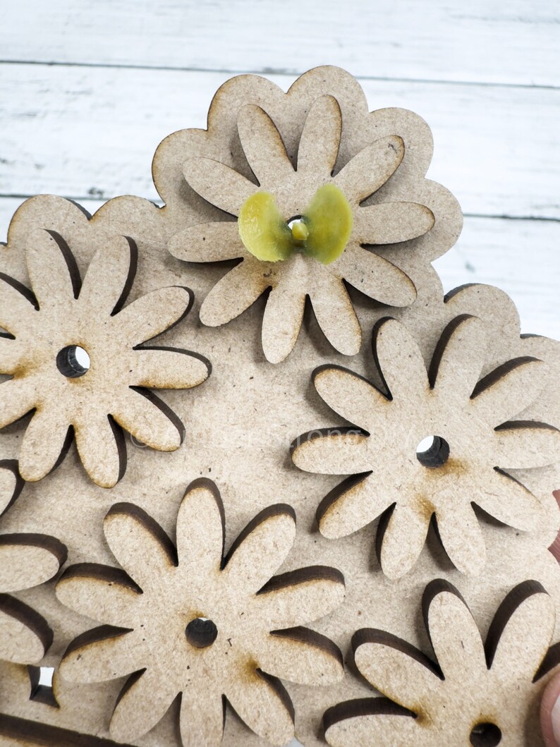 May include: A wooden craft project featuring a design of multiple overlapping flowers. The flowers are cut out of wood and have a brown color. A small yellow flower is placed in the center of the design.