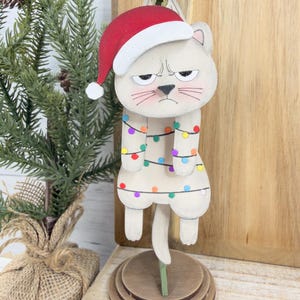 May include: A wooden cat ornament with a Santa hat and string lights. The cat has an annoyed expression and is standing on a wooden base. The ornament is painted in neutral tones with colorful accents, perfect for holiday decor.