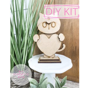 May include: DIY kit featuring a wooden cutout of a cat holding a heart. The cat is light brown and has a white face with black eyes and a black nose. The heart is also light brown. The kit is labeled "DIY Kit".