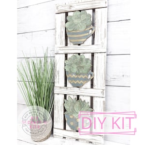 May include: DIY kit for a three-tiered succulent wall decor. The kit includes three wooden cups with painted succulent designs, a whitewashed wooden pallet, and the text "DIY KIT".
