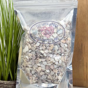 May include: A clear, silver-sealed bag filled with small, multi-colored decorative stones. The bag has a label with the text "DIY WITH AMBER THE MAKERS MAP" in a circular design. The stones are primarily beige, white, and gray.