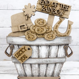 May include: Wooden craft kit featuring a pot of gold at the end of the rainbow. The kit includes a wooden bucket, a horseshoe, a leprechaun hat, a shamrock, gold coins, and a tag that says "Lucky You".