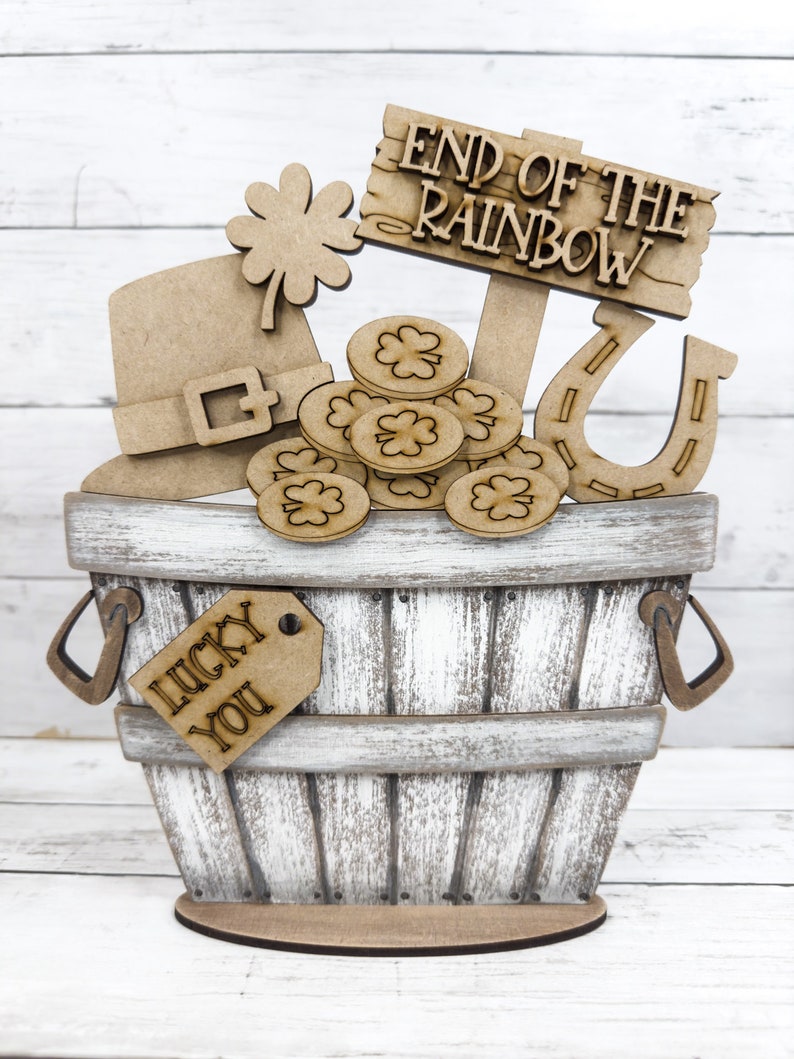 May include: Wooden craft kit featuring a pot of gold at the end of the rainbow. The kit includes a wooden bucket, a horseshoe, a leprechaun hat, a shamrock, gold coins, and a tag that says "Lucky You".