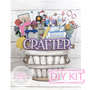 May include: A DIY craft kit featuring a wooden basket overflowing with colorful craft supplies, including paint, yarn, scissors, buttons, and a pencil. The basket is labeled "CRAFTER" and the kit is labeled "DIY KIT".