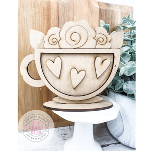 Op de afbeelding: Houten uitsnijding van een koffiekopje met een swirl-ontwerp aan de bovenkant en harten aan de zijkant. Het kopje staat op een kleine houten standaard. De woorden "DIY CRAFT KITS THE MAKERS MAP" staan op een roze cirkel.