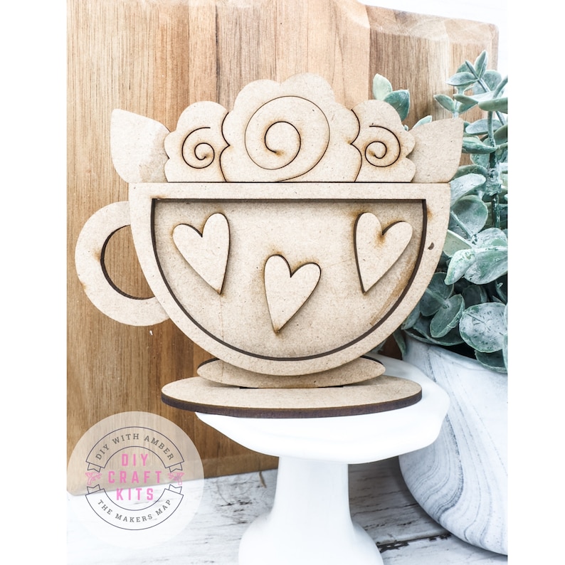 Op de afbeelding: Houten uitsnijding van een koffiekopje met een swirl-ontwerp aan de bovenkant en harten aan de zijkant. Het kopje staat op een kleine houten standaard. De woorden "DIY CRAFT KITS THE MAKERS MAP" staan op een roze cirkel.