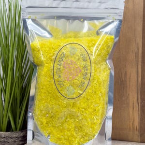 May include: A clear, sealed bag filled with yellow, granular material. The bag has a silver top and a circular label with the text "DIY WITH AMBER" and "THE MAKERS MAP". The background includes green plants and a wooden surface.